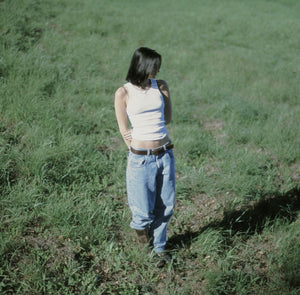 Person standing in a grassy field wearing a white tank top and blue jeans.